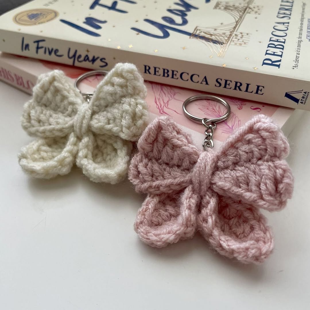 Two crocheted butterfly keychains, one white and one pink, on a surface with books in the background.
